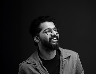 Black-and-white studio portrait of Manvith, featuring a candid side-facing pose with glasses and a layered jacket for the JUMPINGGOOSE team page.