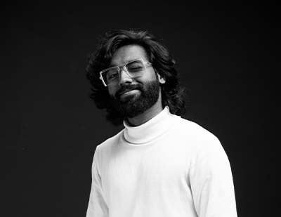 Black-and-white studio portrait of Mashood, The Animatron, featuring a relaxed pose in a high-neck sweater alongside a bold title card highlighting his creative role at JUMPINGGOOSE.