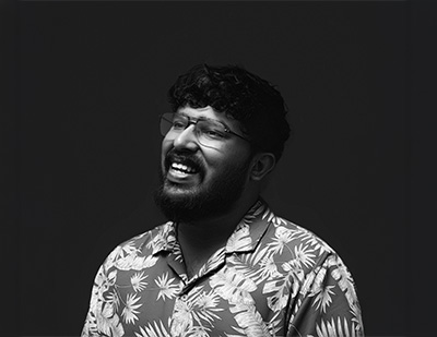 Black-and-white studio portrait of Midhun, featuring a cheerful side-facing pose with glasses and a patterned shirt for the JUMPINGGOOSE team page.