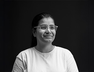 Black-and-white studio portrait of Nitika, featuring a soft side-facing smile with glasses and a simple white T-shirt for the JUMPINGGOOSE team page.